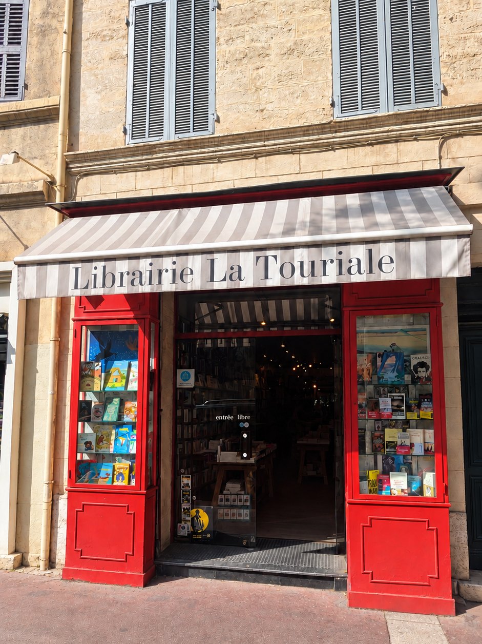 La devanture de la librairie la Touriale au soleil, boiseries rouges, store rayé et vitrines pleines de livres
